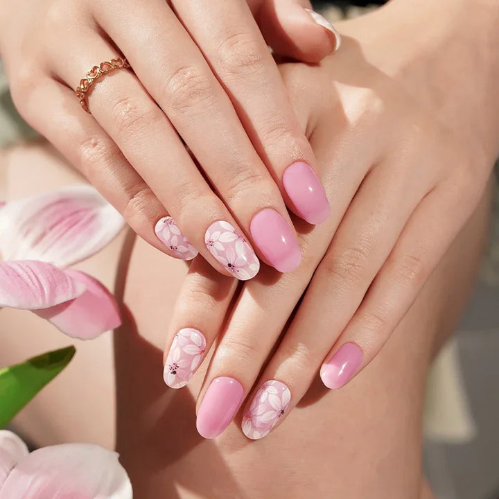 Semi-cured gel nail wraps in blush pink with white floral patterns and gold accents, featuring a translucent gloss finish, shown on fair skin wearing gold rings.