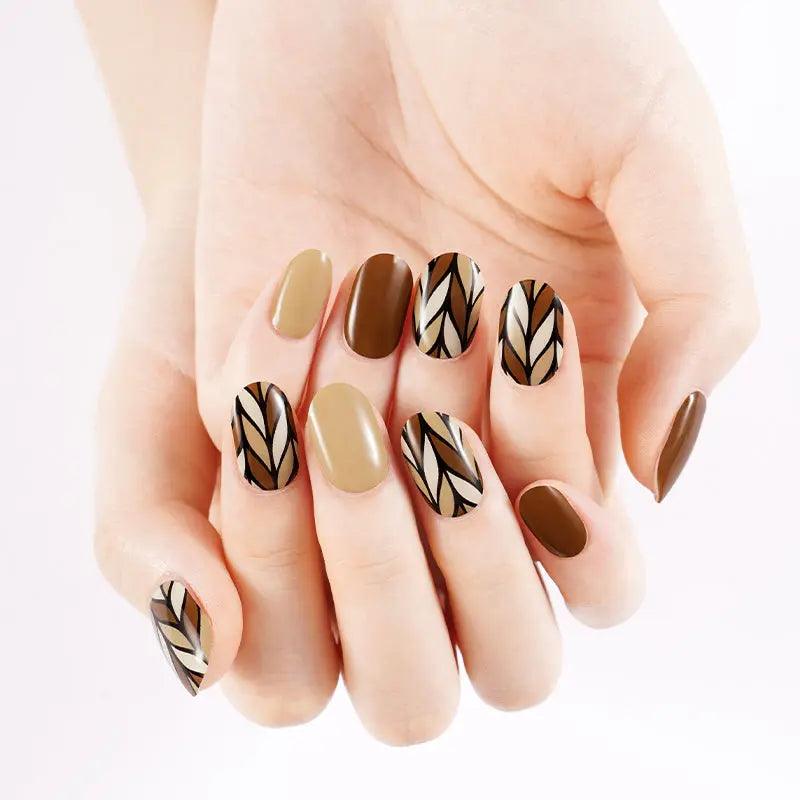 Caramel and nude gel nail wraps with black and beige geometric leaf accents, glossy finish on light skin tone under bright studio light.