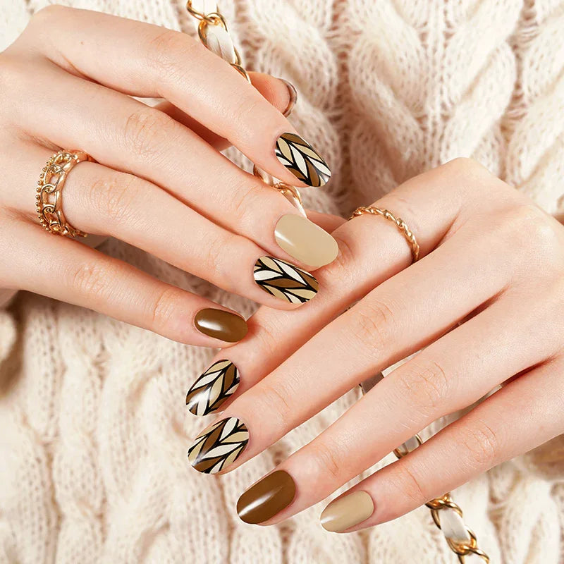 Caramel and nude gel nail wraps with black geometric accents and glossy finish on light skin tone, styled with gold rings and cream knit sweater.