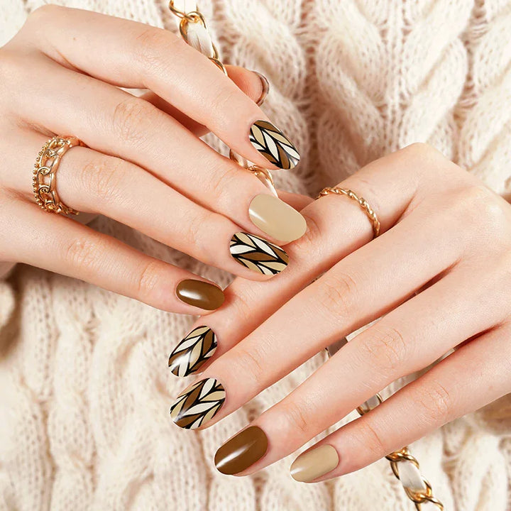 Caramel and nude gel nail wraps with black geometric accents and glossy finish on light skin tone, styled with gold rings and cream knit sweater.