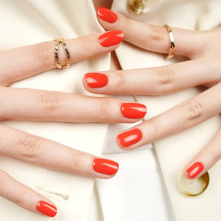Semi-cured gel nail wraps in bright watermelon red with an orange undertone and high-gloss finish, shown on fair skin wearing gold jewelry and a cream outfit.