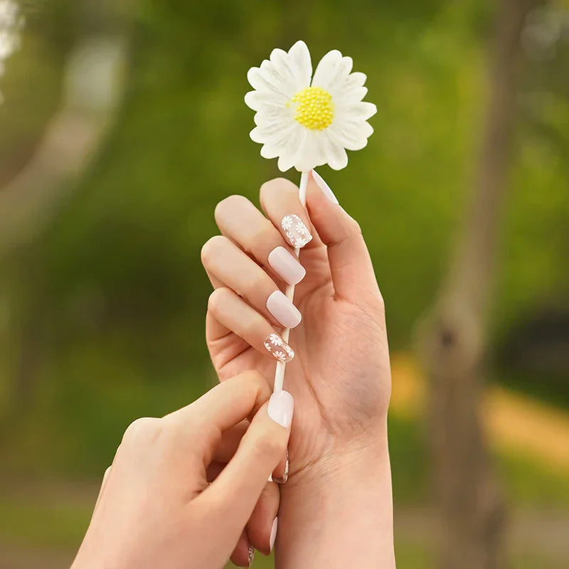 Baby pink gel nail wraps with glossy finish and white daisy accents on light skin tone, photographed outdoors.