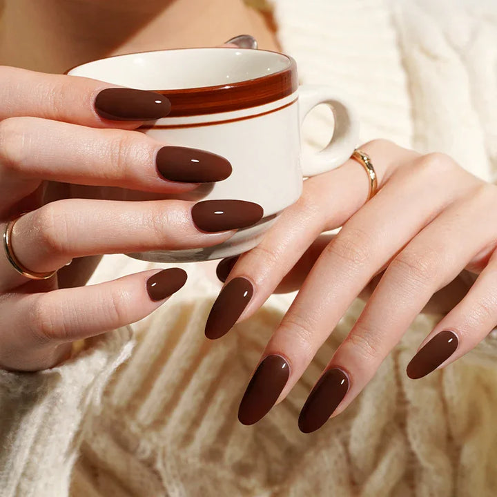 Deep coffee brown gel nail wraps with glossy finish on light skin tone, styled with gold rings and cream knit in warm indoor light.
