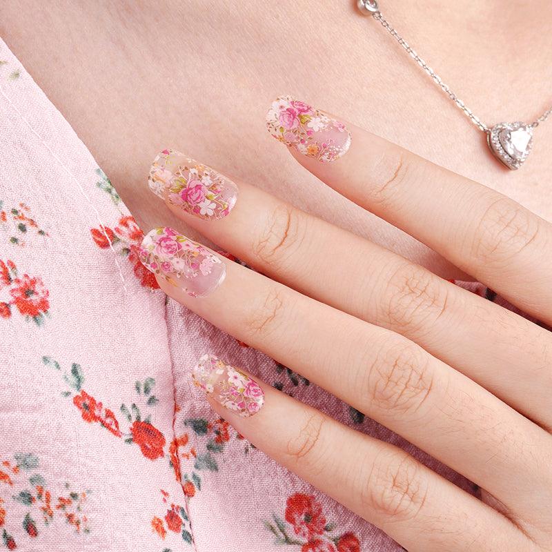 Sheer floral gel nail wraps with transparent base and soft pink, coral, and green flower accents on light skin tone, styled with floral fabric under natural daylight.