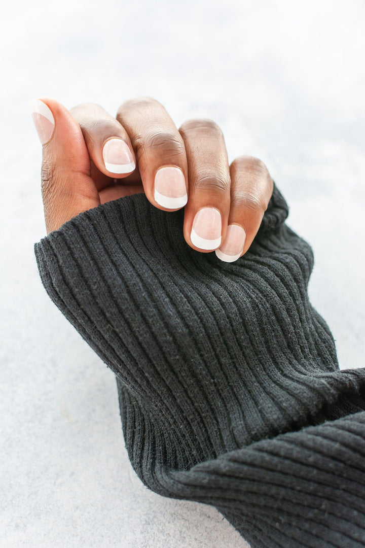 French tip gel nail wraps with soft beige base and white tips on dark skin tone, styled with ribbed grey knit sweater in natural light.