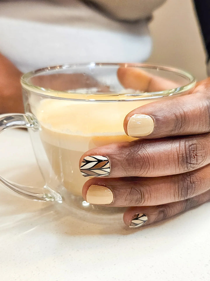 Caramel and nude geometric gel nail wraps with glossy finish on medium skin tone, styled with glass coffee mug in natural indoor light.