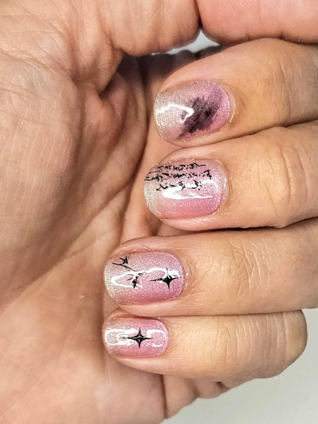 Semi-cured gel nail wraps in sheer pink with a soft cat-eye shimmer, shown on medium-deep skin with glossy finish and black inky nail art details.