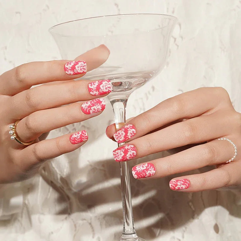 Semi-cured gel nail wraps in vivid pink with white floral hibiscus motifs and a glossy shine, shown on fair skin wearing rings.