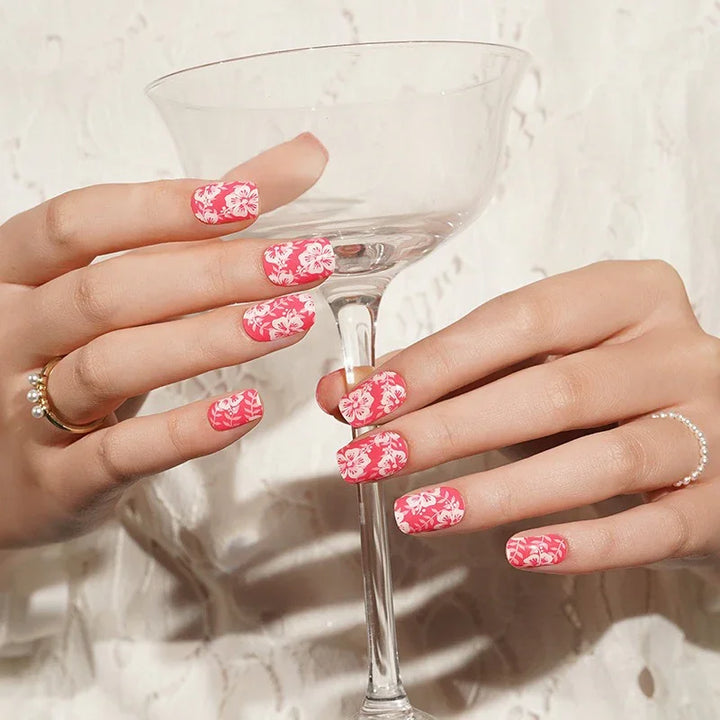 Semi-cured gel nail wraps in vivid pink with white floral hibiscus motifs and a glossy shine, shown on fair skin wearing rings.
