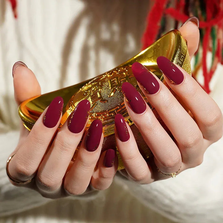 Semi-cured gel nail wraps in deep maroon with gold leaf accents and a high-gloss finish, shown on fair skin wearing a cream knit sweater.