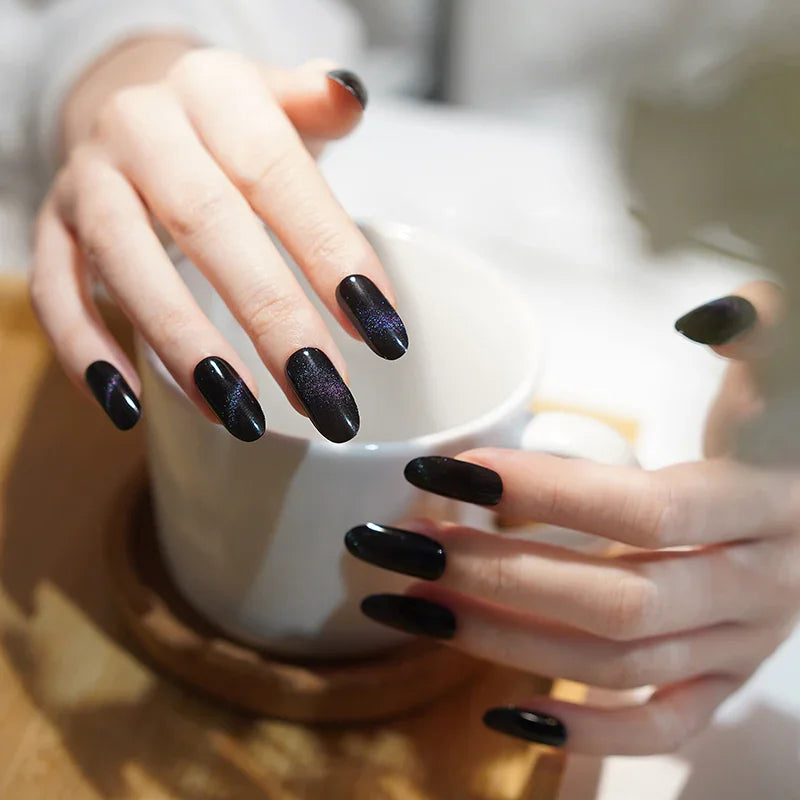 Black cat-eye gel nail wraps with subtle translucency and rainbow shimmer on light skin tone, glossy magnetic finish in soft daylight.