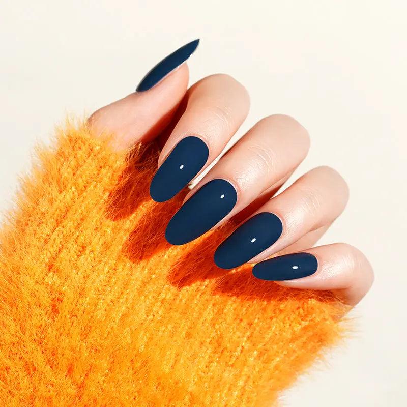 Dark navy blue gel nail wraps with cool undertone and high-gloss finish on light skin tone, styled with bright orange knit sweater.