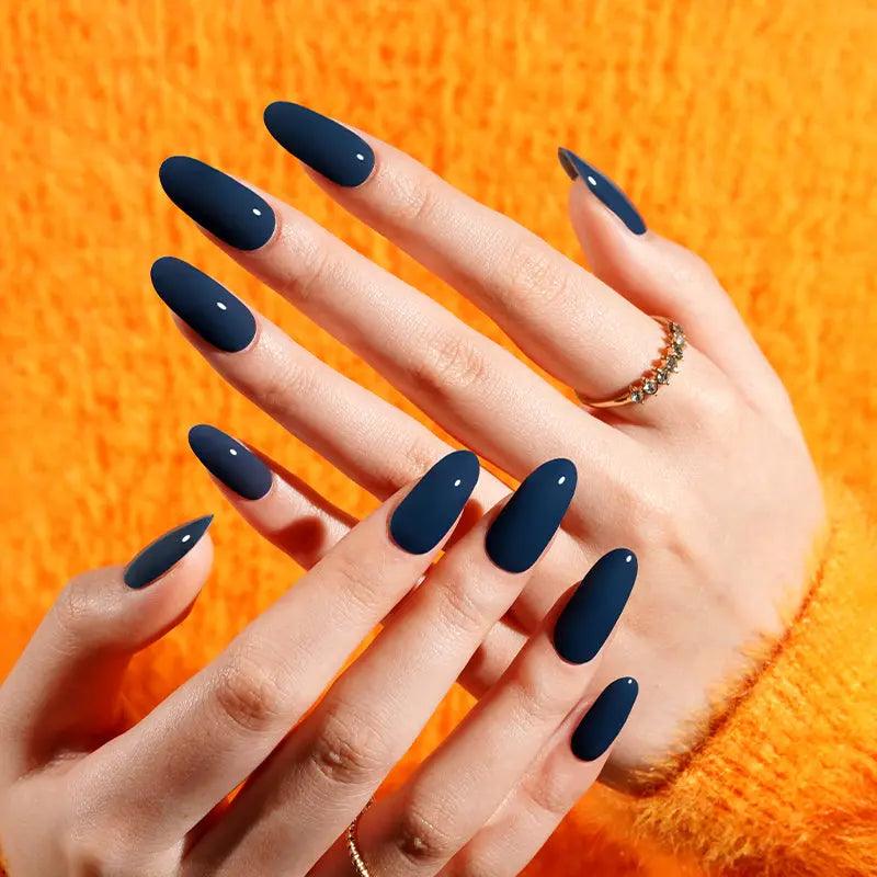 Dark navy blue gel nail wraps with high-gloss finish on light skin tone, styled with gold rings and bright orange knit sweater in natural light.