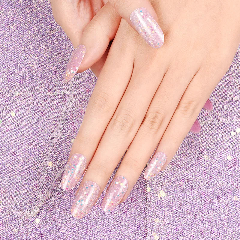 Hand with pink gel nail wraps featuring iridescent glitter and glow-in-the-dark sparkles shown on a purple glitter fabric background.