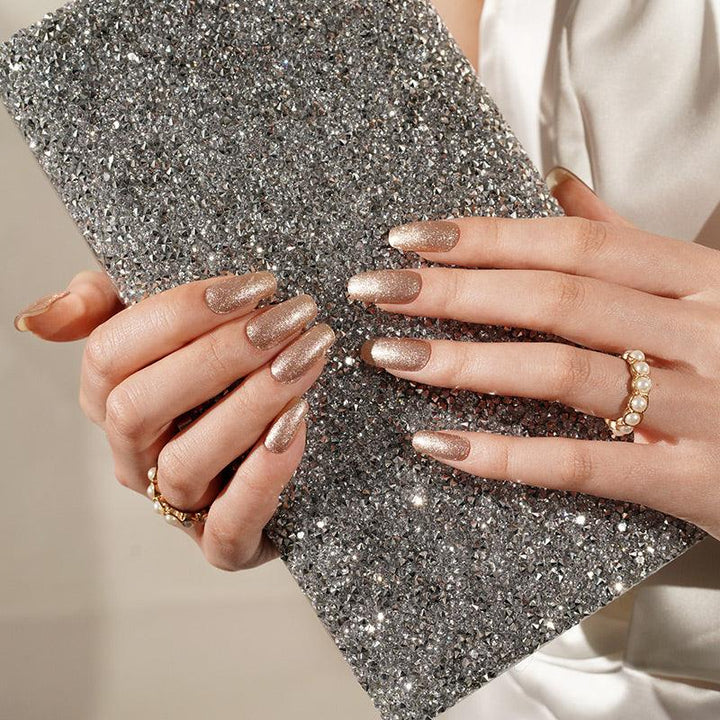 Metallic champagne gel nail wraps with dense shimmer on light skin tone, styled with glitter clutch in warm indoor light.