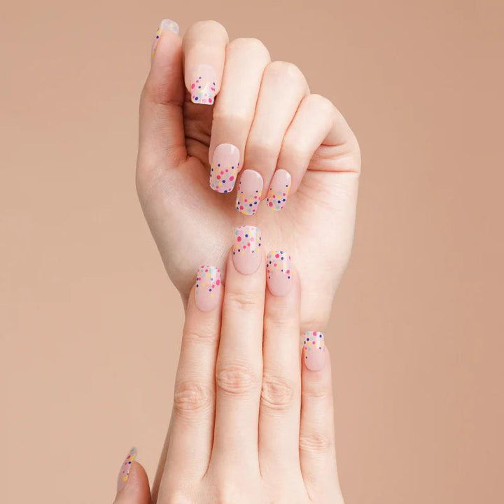Warm nude gel nail wraps with glossy finish and scattered rainbow polka dots on light skin tone under soft lighting.