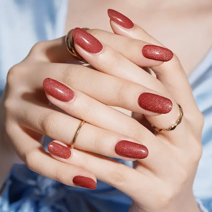 Semi-cured gel nail wraps in brick-red with subtle gold shimmer and glossy cat-eye depth, shown on light-medium skin wearing gold rings and blue fabric.