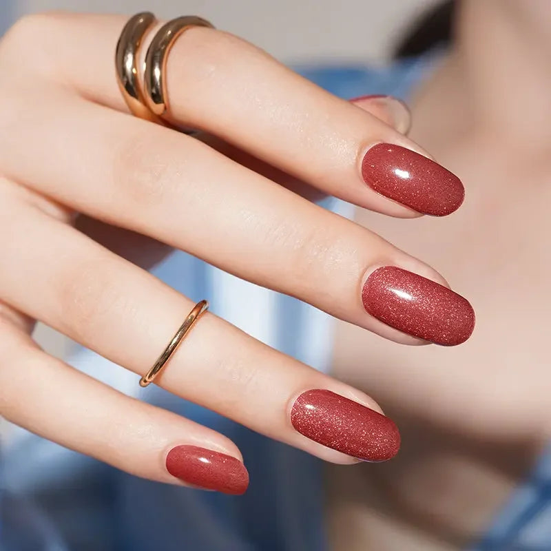 Semi-cured gel nail wraps in rich rust-red with magnetic shimmer and high-gloss reflection, shown on light skin wearing gold jewelry.