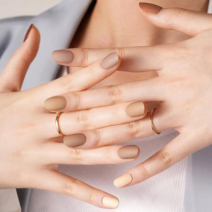 Matte neutral gel nail wraps in sandy beige and brown tones on light skin tone, styled with gold rings in soft studio light.