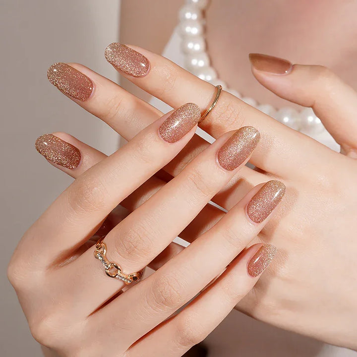 Semi-cured gel nail wraps in sheer gold with fine shimmer and a luminous cat-eye glow, shown on light skin wearing gold rings and a pearl necklace under warm studio lighting.