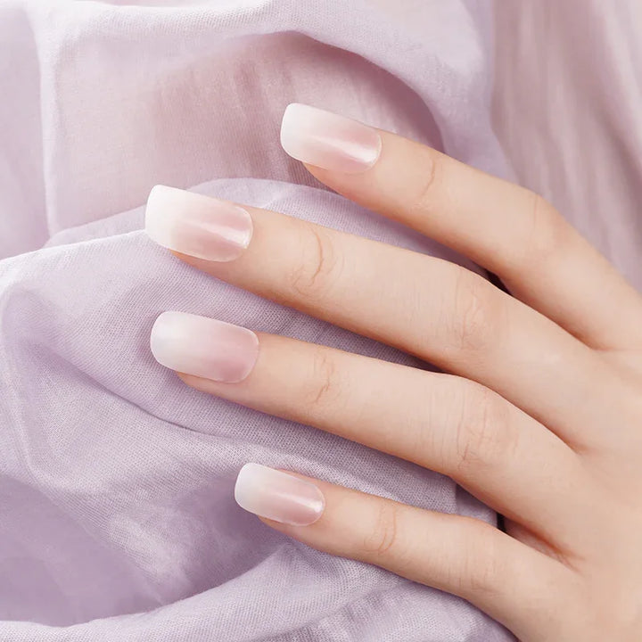 Semi-transparent nude ombré gel nail wraps with glossy finish on light skin tone, photographed against soft lilac fabric.