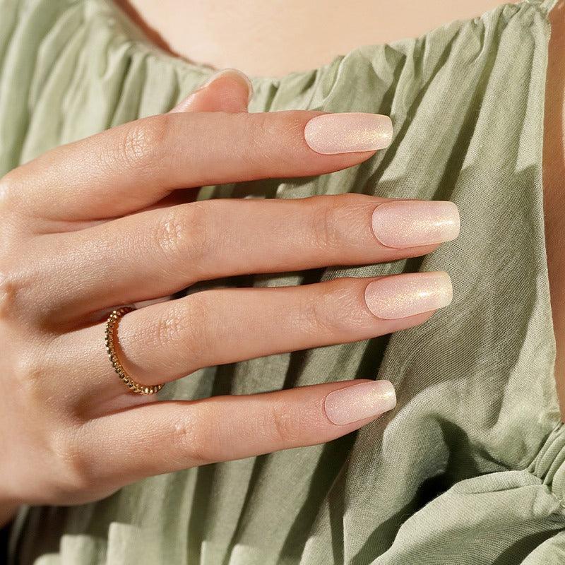 Semi-cured gel nail wraps in milky nude with an ultra-fine shimmer that shifts from gold to silver under light, shown on fair skin wearing a gold ring and sage-green fabric.