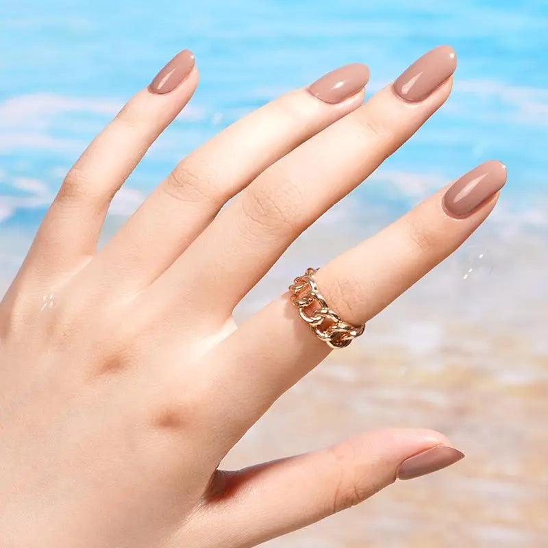 Glossy taupe gel nail wraps with creamy rose-brown undertone on light skin tone, photographed in bright daylight near blue water.