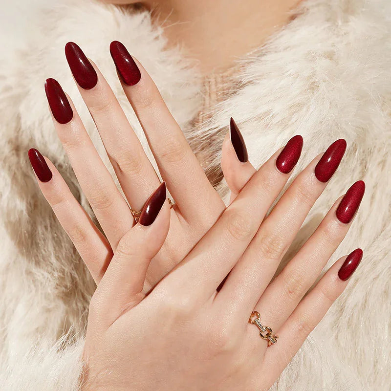 Semi-cured gel nail wraps in deep wine red with black undertones and a magnetic cat-eye shimmer, shown on light skin wearing gold rings and a soft faux-fur coat.