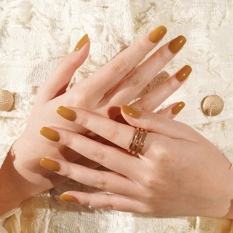 Light-skin hands wearing glossy burnt-caramel semi-cured gel nail stickers under soft studio lighting, paired with a gold ring and a textured cream jacket.