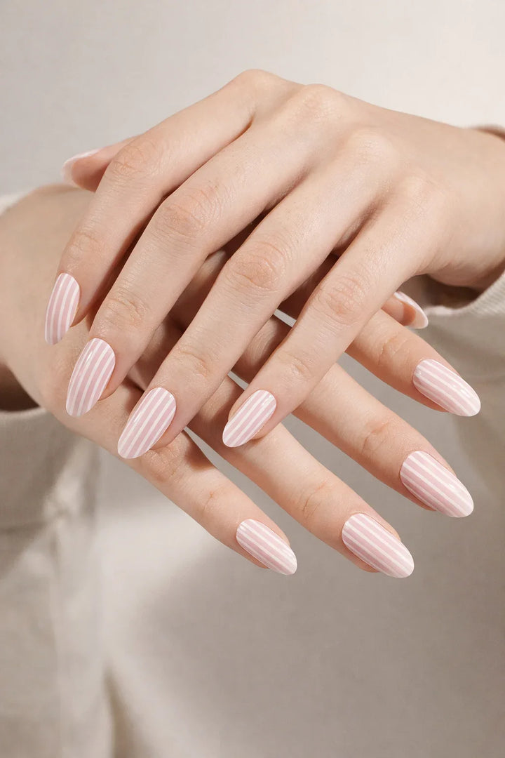 Candy Stripe semi-cured gel nail stickers, shown on light skin with a semi-sheer pale pink base and fine vertical white stripes, finished with a smooth high-gloss surface, photographed under soft studio lighting against a neutral beige background.