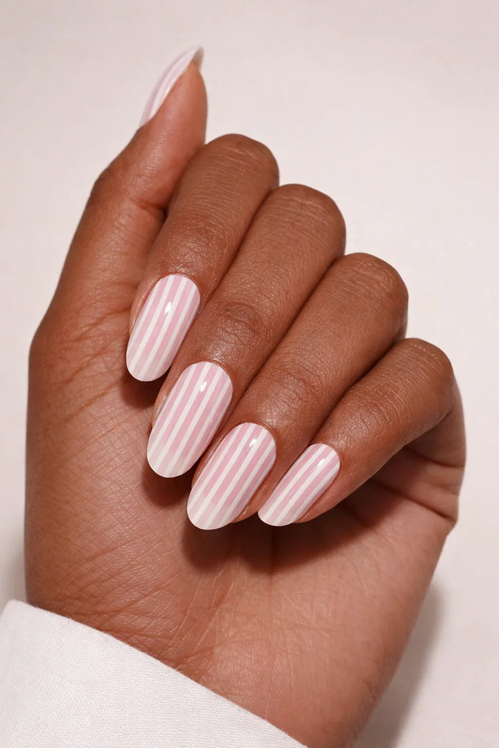 Candy Stripe semi-cured gel nail stickers, shown on medium-deep brown skin with a semi-sheer soft pink base and fine vertical white stripes, finished in a smooth high-gloss, photographed under soft studio lighting against a clean white background.