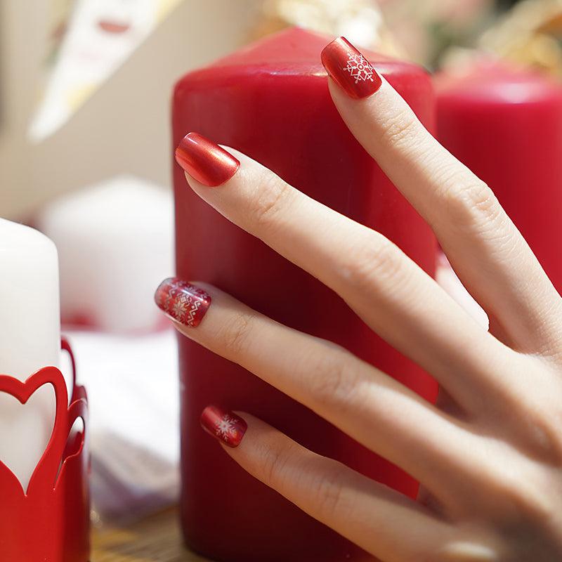 Red gel nail wraps with white snowflake accents on light skin tone, styled with red candle in soft lighting.
