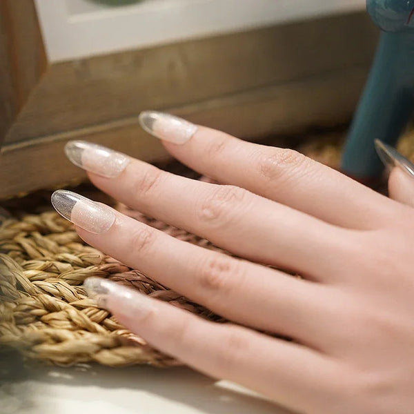 Hands with Clear Cat Eye gel nail wraps featuring a soft shimmer and translucent glow, shown resting on a woven mat in natural light.