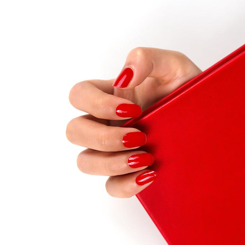 Light-skinned hand with crimson red gel nail wraps in a high-gloss finish against a white background.