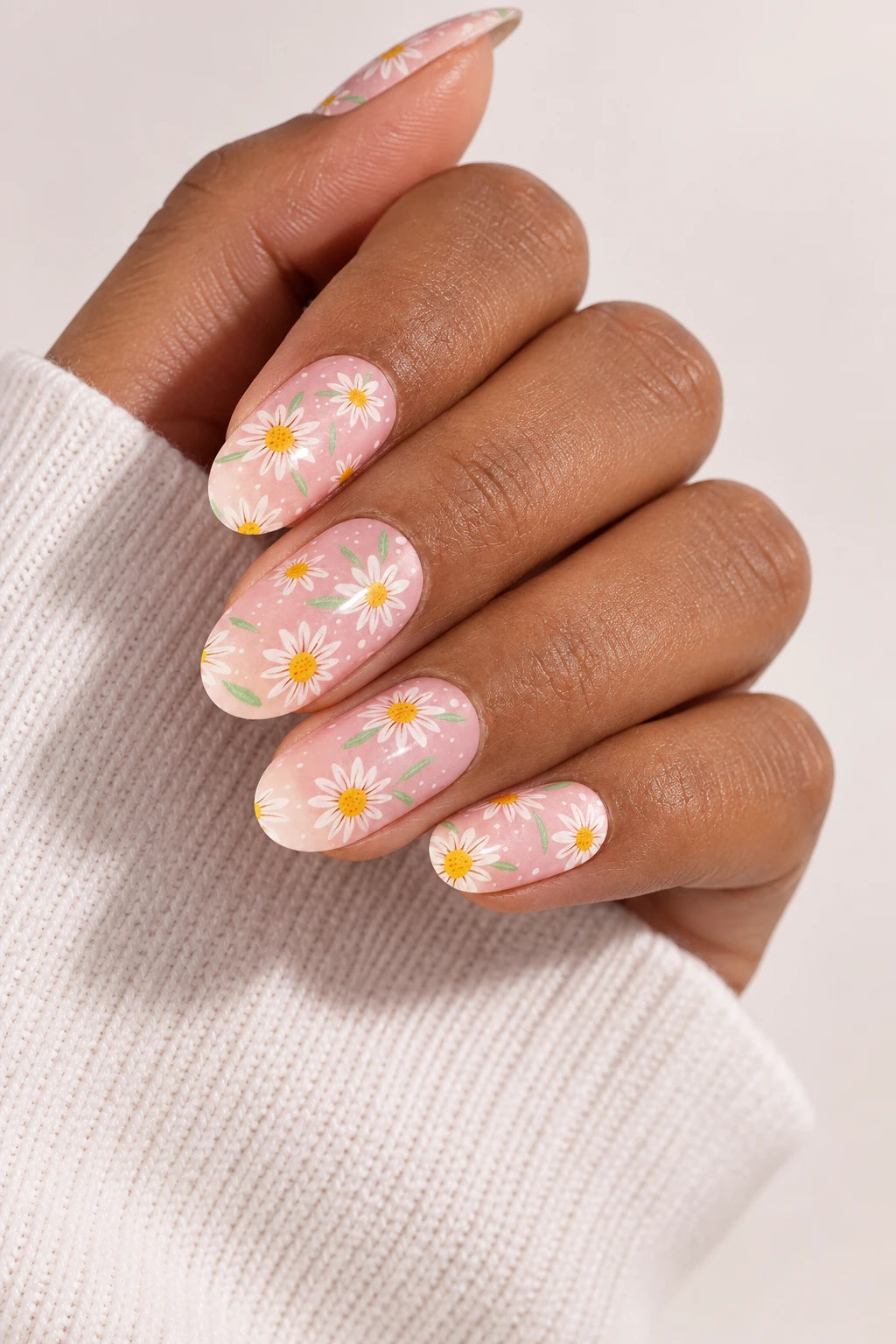 UV-free gel nail wraps featuring the Daisy Daze design with white daisies on a soft pink base, shown on medium-brown skin with cream knit clothing.