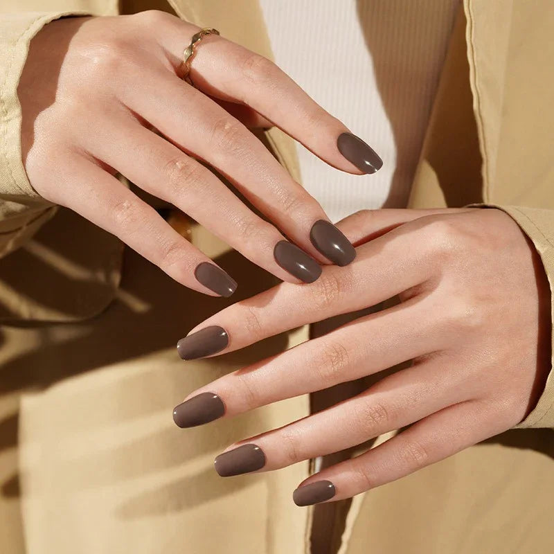 Dark Truffle semi-cured gel nail stickers in a deep taupe-brown shade with a smooth glossy finish, shown under warm studio lighting on light skin with gold rings and a beige jacket.