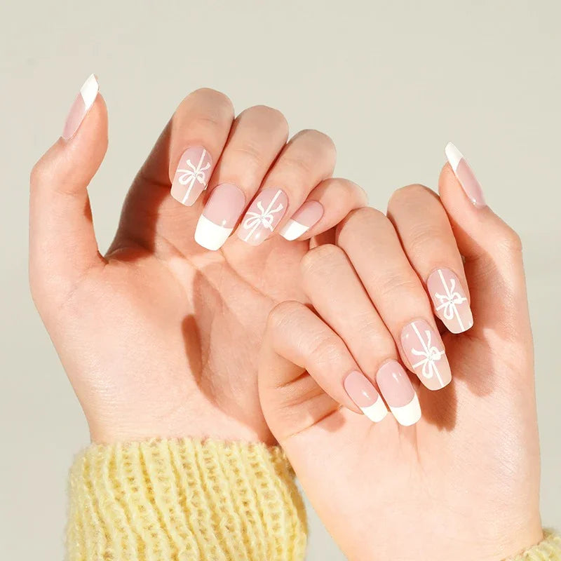 Hands in a yellow knit sweater showing French tip nails with tiny white bows on a soft pink base, photographed in bright neutral light.