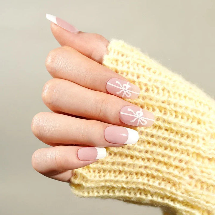 Hands wrapped in a yellow knit sweater displaying French tip nails with white bow accents, captured in soft natural lighting.