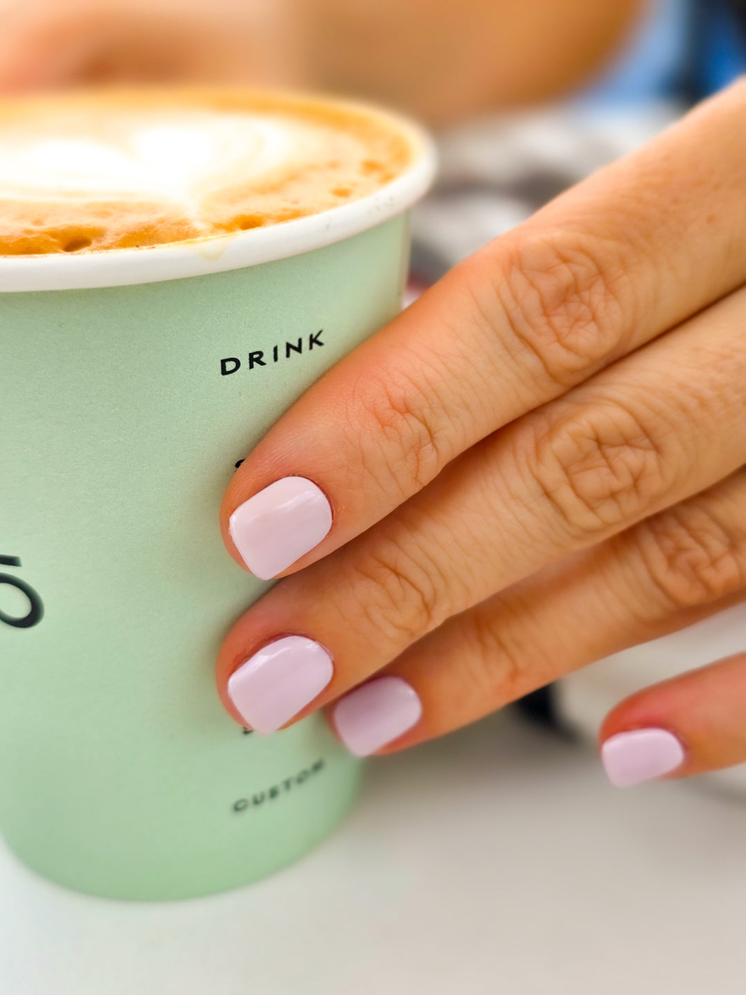 Hand holding a green cup with a cappuccino, wearing pink nail polish.