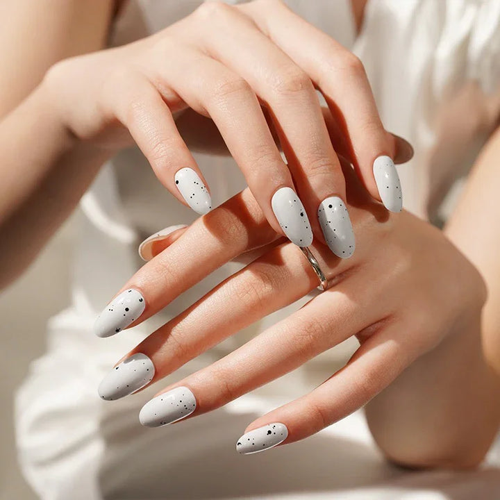 Hands wearing Gray Speckled Eggs gel nail wraps in glossy pale grey with delicate black flecks, shown in soft natural light against a neutral background.