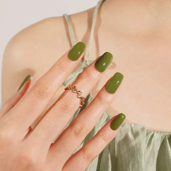Semi-cured gel nail wraps in deep olive green with a bold, glossy finish, shown on light-medium skin wearing gold rings and a sage satin dress.