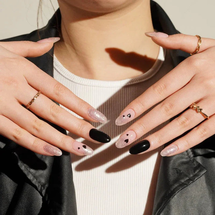 Hands wearing It Girl gel nail stickers in a glossy pink-nude base with black heart nail art, shimmer tips, and bold black accent nails.
