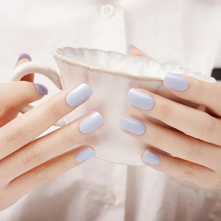 Semi-cured gel nail wraps in pastel periwinkle with a glossy sheen, shown on light skin while holding a ceramic cup in natural indoor light.