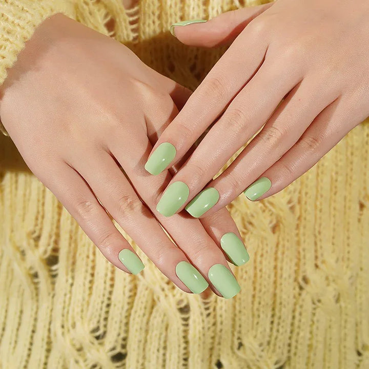 Light-skin hands wearing glossy pistachio-green semi-cured gel nail stickers, shown under soft studio lighting against a yellow knitted sweater.