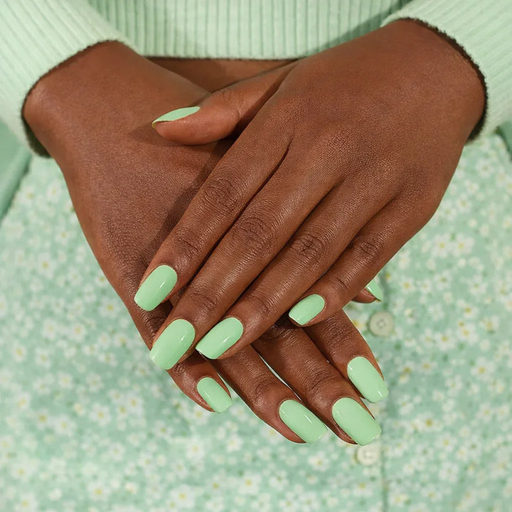 Dark-medium skin hands wearing glossy pistachio-green semi-cured gel nail stickers, photographed under soft studio lighting against a mint-green outfit with a subtle floral pattern.