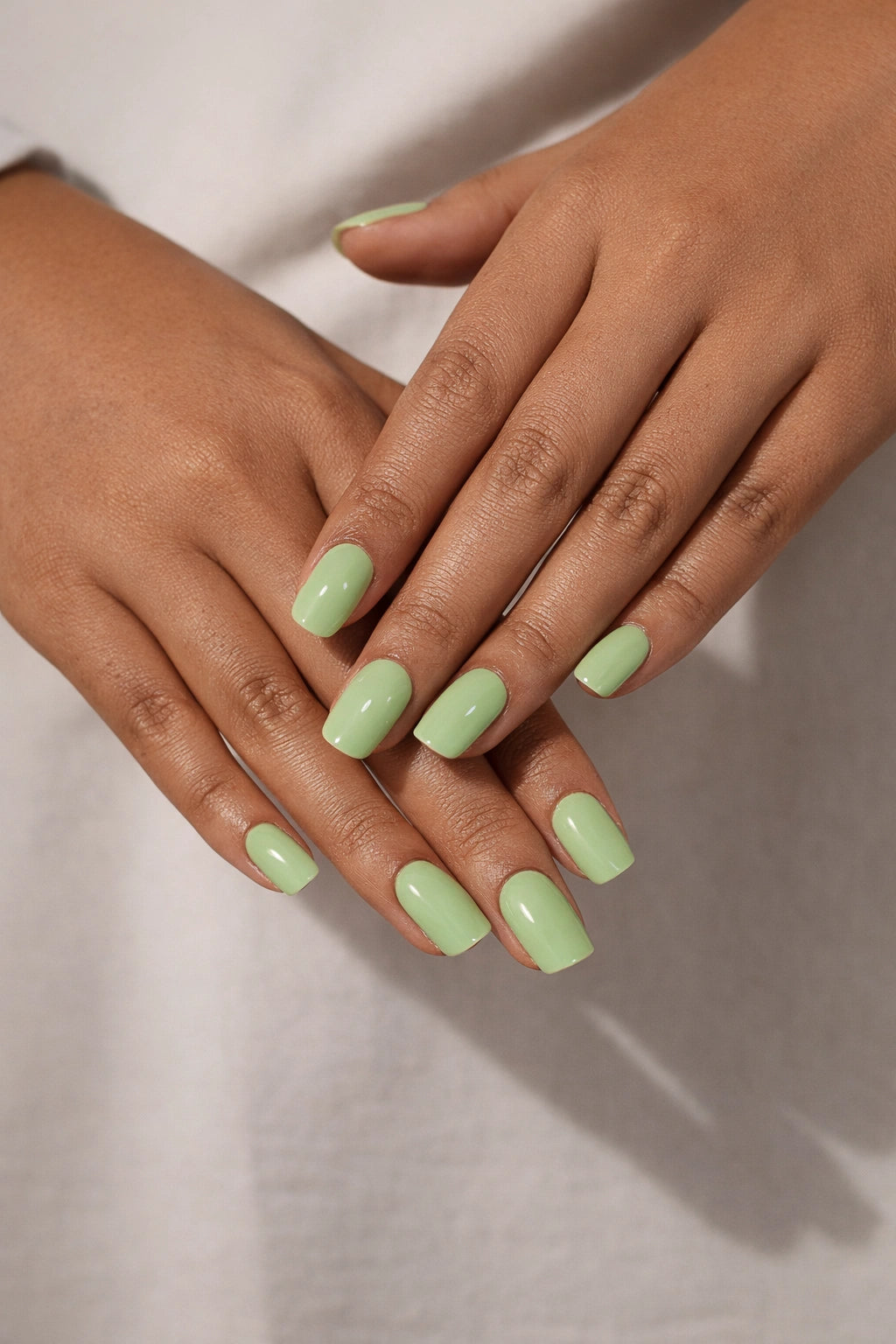 Pistachio semi-cured gel nail stickers, worn on medium brown skin with a creamy pastel green tone and glossy finish, photographed under soft studio lighting against a neutral fabric backdrop.