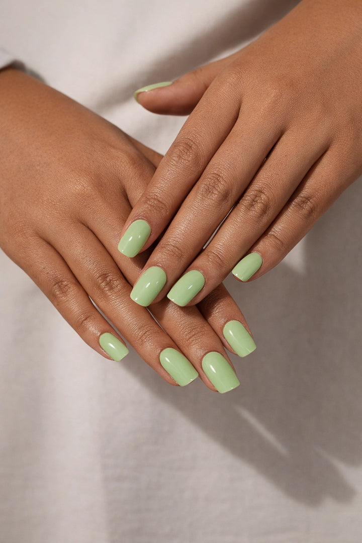 Pistachio semi-cured gel nail stickers, worn on medium brown skin with a creamy pastel green tone and glossy finish, photographed under soft studio lighting against a neutral fabric backdrop.