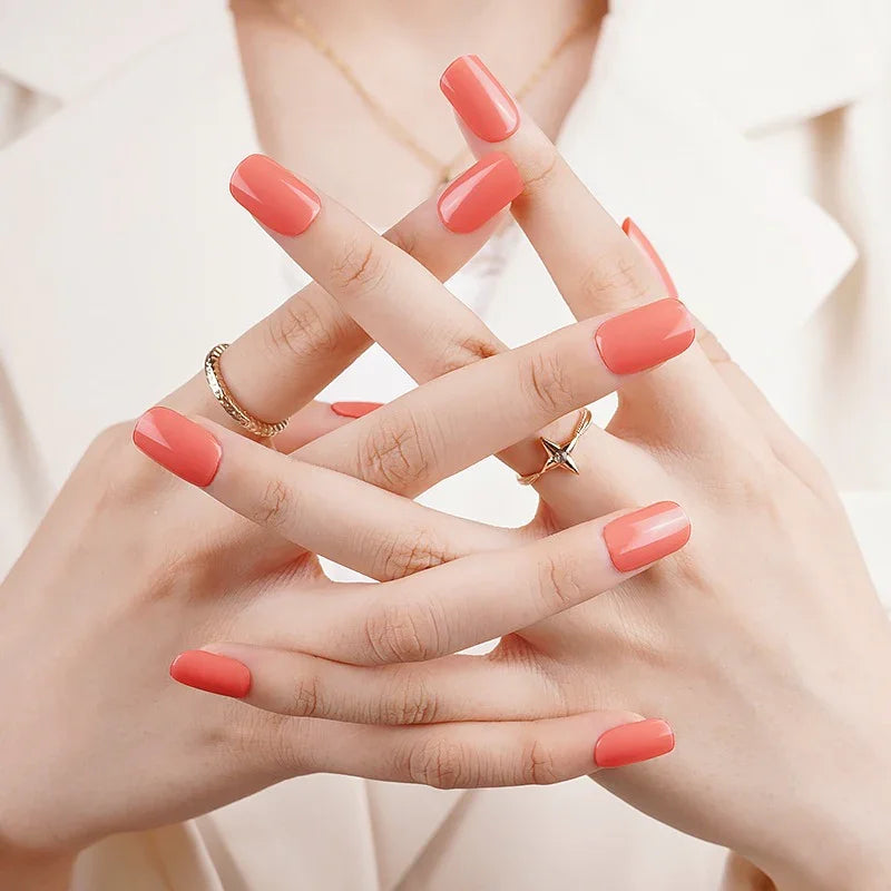 Semi-cured gel nail wraps in bold pink-red coral with a glossy shine, shown on fair skin wearing a cream outfit