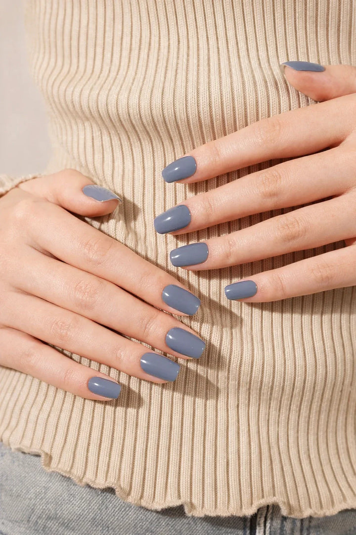 Slate Blue semi-cured gel nail stickers in a muted blue-grey shade with a glossy finish, shown under soft studio lighting on light skin with a beige ribbed top.