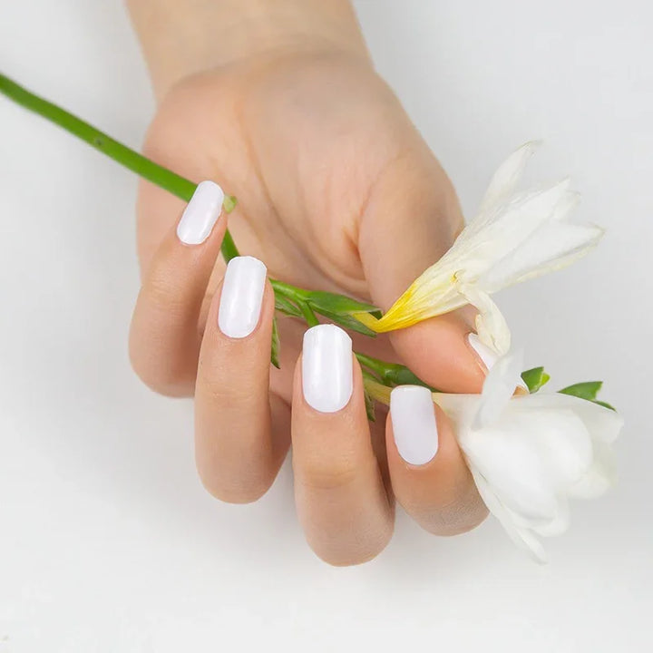 Semi-cured white gel nail wraps with a shiny, icy gloss on fair skin, shown holding a white flower in soft natural light.